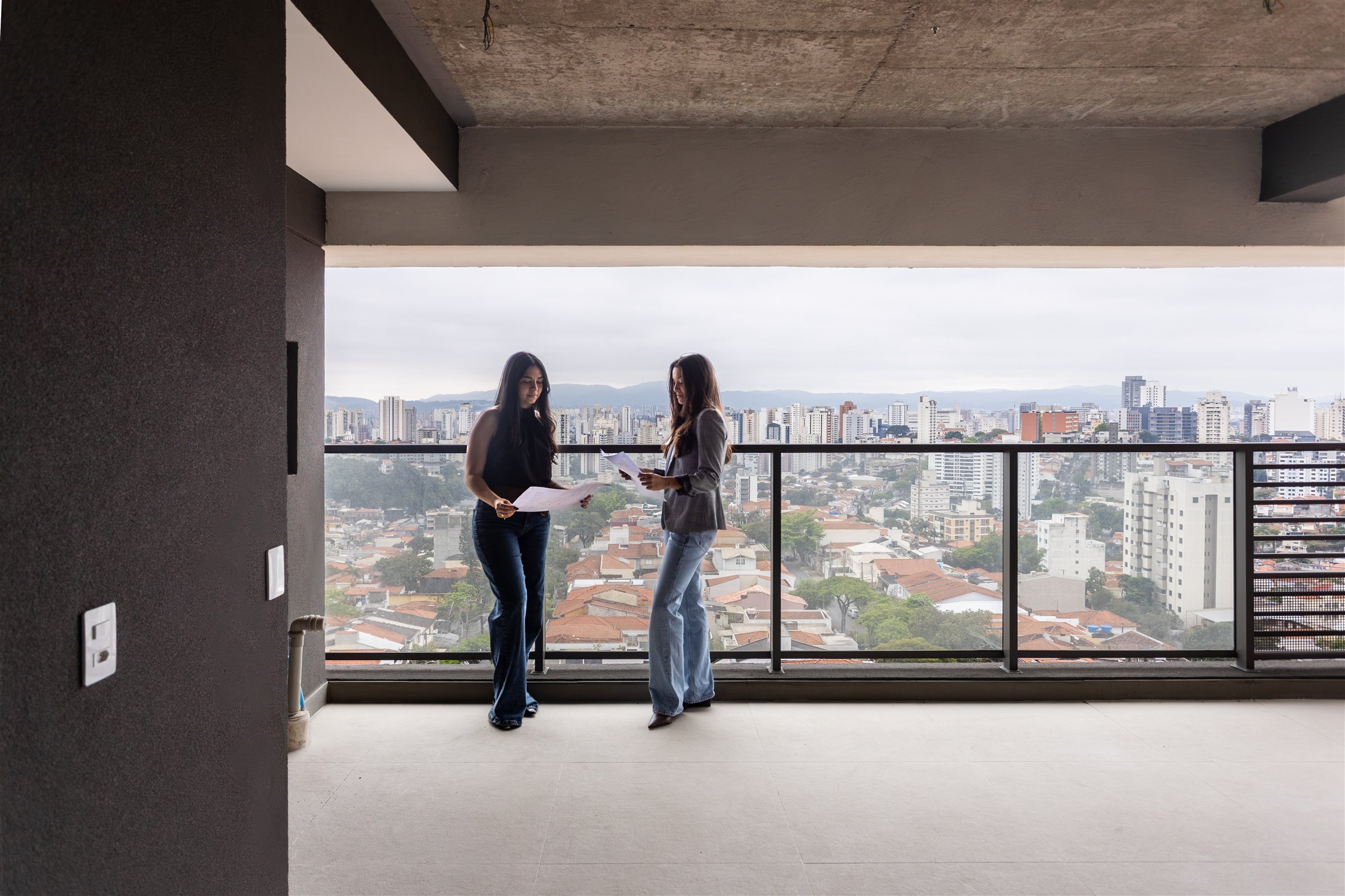 Marianna e Marina na varanda de um apartamento em obra, olhando as plantas do projeto